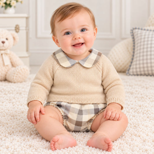 A smiling baby sitting on a white shag rug wearing a beige knit sweater with a plaid Peter Pan collar and matching plaid diaper cover, with a teddy bear and soft room decor in the background.