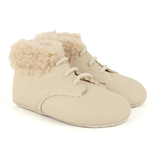 A pair of cream-colored baby booties featuring a smooth faux-leather upper, decorative laces, and a fluffy cream sherpa lining at the ankle opening, shown on a white background.
