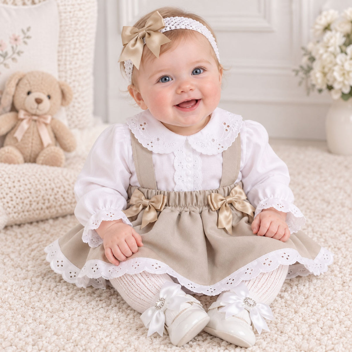 A baby girl sitting on a cream carpet wearing a two-piece dress set consisting of a white long-sleeve blouse with a lace Peter Pan collar and a beige pinafore-style skirt with matching beige bows, a beige headband with a bow, and white lace-up baby shoes.