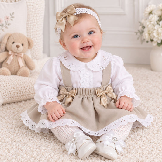 A baby girl sitting on a cream carpet wearing a two-piece dress set consisting of a white long-sleeve blouse with a lace Peter Pan collar and a beige pinafore-style skirt with matching beige bows, a beige headband with a bow, and white lace-up baby shoes.