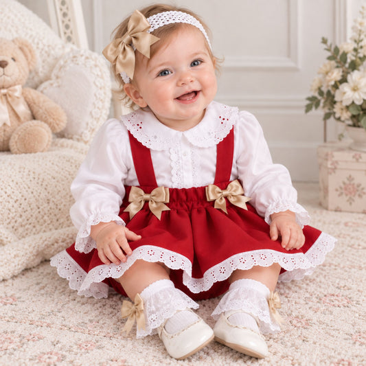 A baby girl wearing a red and white holiday-themed outfit with beige bows and a matching headband, sitting on a floral rug in a cozy room setting.