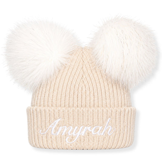 Beige knit beanie with white fur pom-poms and personalised name embroidery on a white background