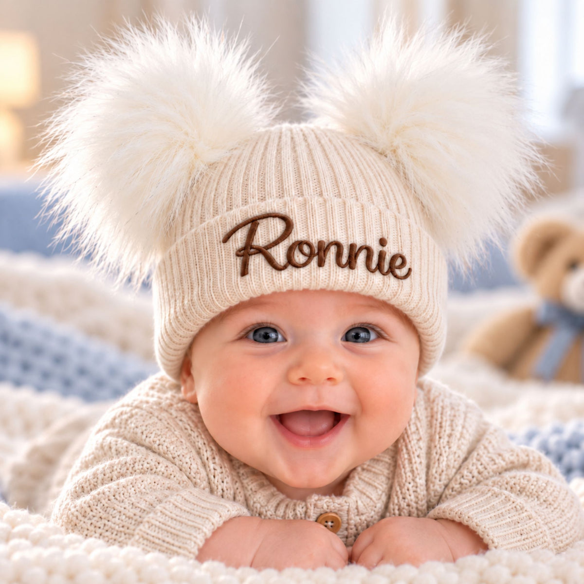 A smiling baby wearing a cream knit beanie with two large fluffy pom-poms and the name 'Ronnie' embroidered in brown on the front, lying on a soft cream blanket.