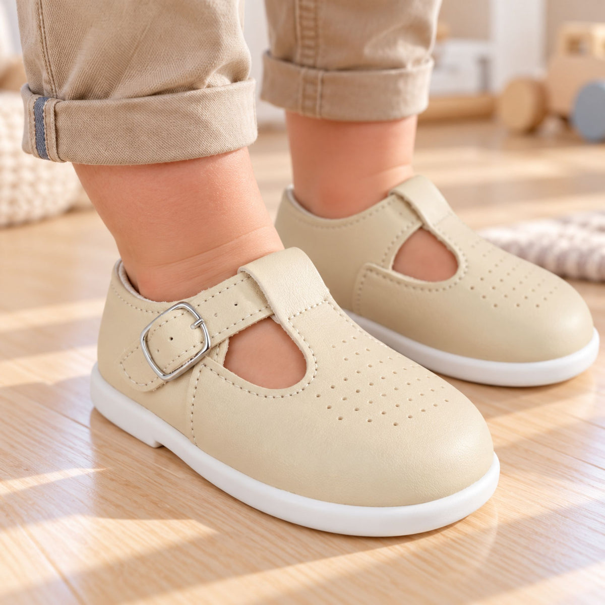 A close-up of a child wearing light beige Mary Jane-style shoes with a hook-and-loop strap and silver buckle, paired with tan pants, standing on a light wood floor.
