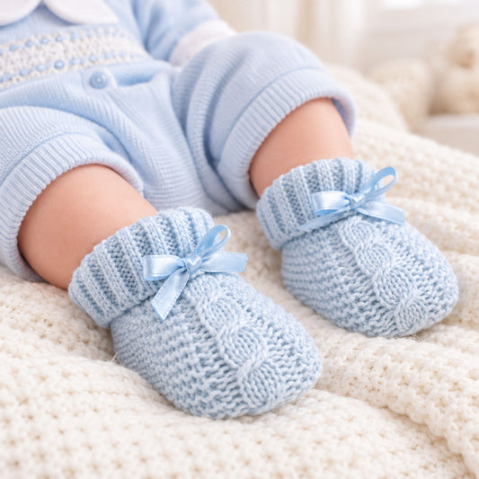 A close-up of a baby's feet wearing light blue cable-knit booties with matching ribbon bows, styled with a light blue knit outfit on a cream knit blanket.