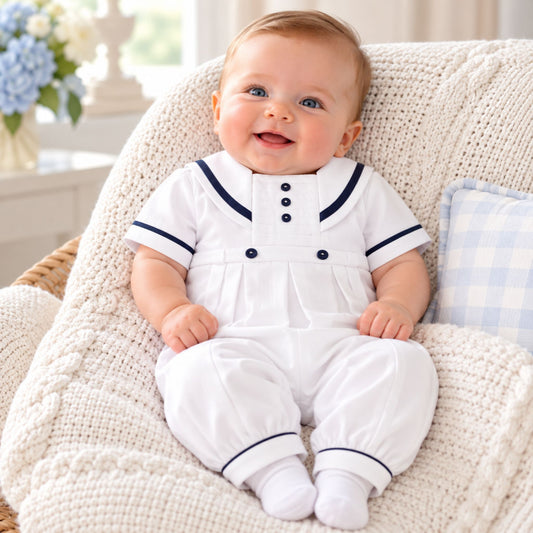 A baby sitting on a cream cable-knit chair wearing a white short-sleeve romper with navy blue trim and buttons, styled with a blue and white gingham pillow in the background.