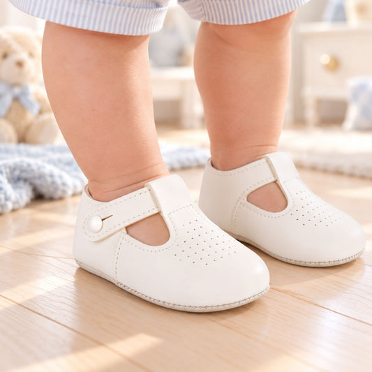 A baby wearing a pair of off-white Mary Jane style baby shoes with a strap and button closure, standing on a light wooden floor in a softly lit nursery.