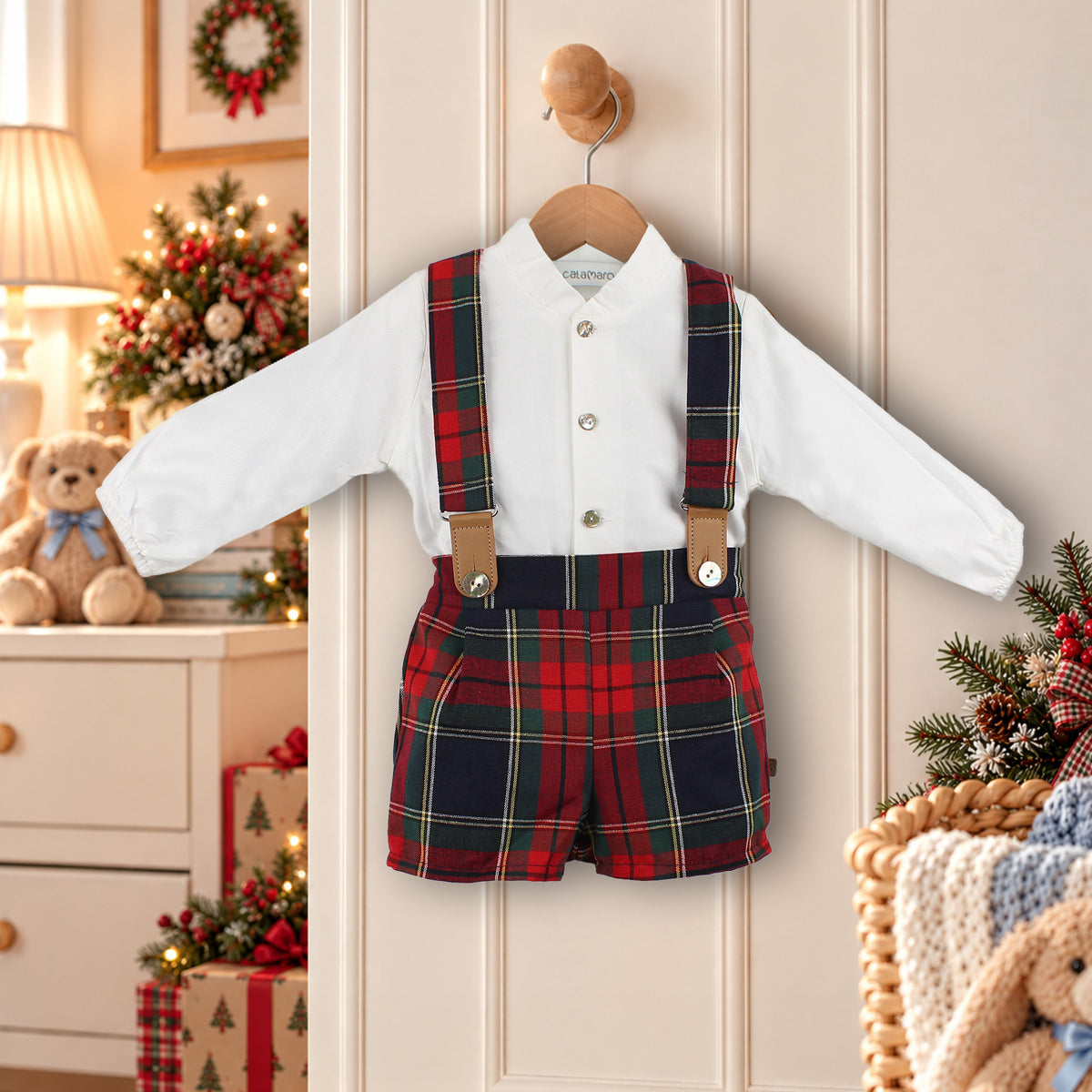 A baby boy's two-piece clothing set featuring a white long-sleeve button-up shirt and red, green, and navy plaid shorts with suspenders, hanging on a wooden hanger against a cream paneled door with a cozy Christmas-themed background.