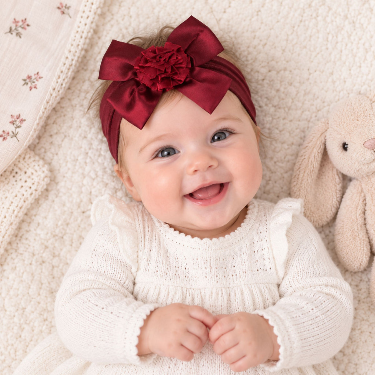 A smiling baby lying on a soft cream blanket, wearing a large burgundy satin bow headband with a flower accent and a white knit dress. A beige stuffed bunny is beside the baby.