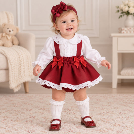 A toddler standing indoors wearing a red and white baby dress outfit with a white blouse, red skirt, matching red bow headband, white knee socks, and red shoes.