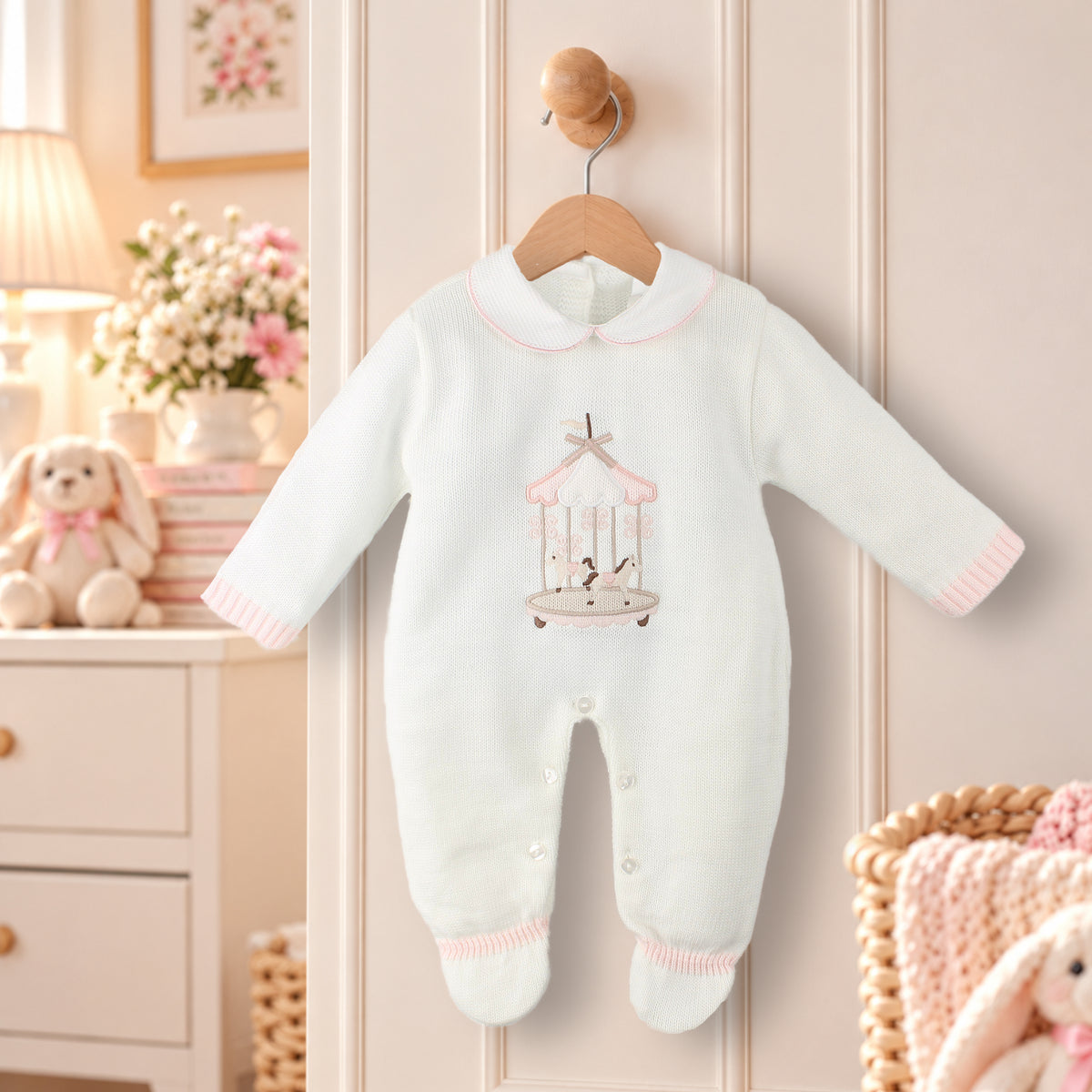 A baby long-sleeve footed romper in white with a pink and brown carousel graphic on the chest, hanging on a wooden hanger against a cream paneled door.
