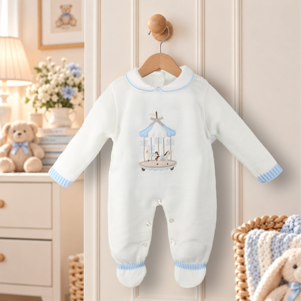 A white long-sleeve baby footed sleeper with a light blue collar, cuffs, and ankle trim, featuring an embroidered carousel design on the chest. The sleeper is hanging on a wooden hanger from a wooden door knob in a cozy, styled nursery setting.