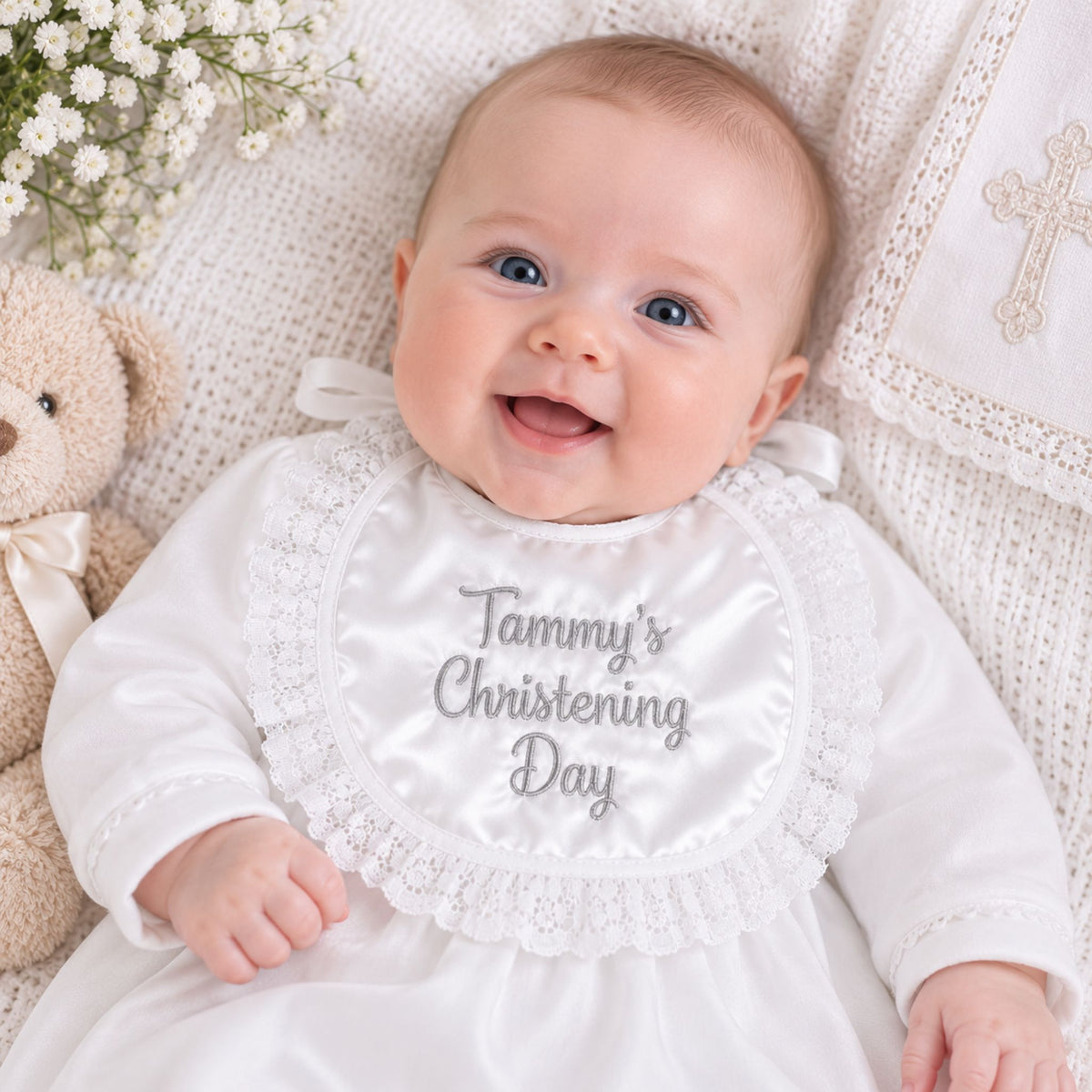 A smiling baby lying on a white knit blanket, wearing a white satin christening gown and bib with silver embroidered text reading 'Tammy's Christening Day.' A teddy bear and small white flowers are visible at the sides.