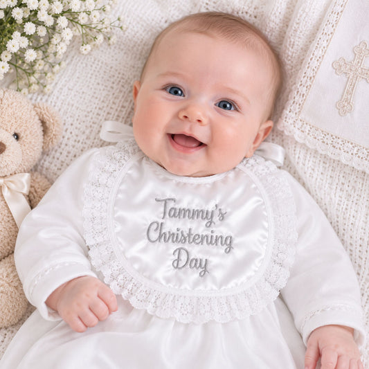 A smiling baby lying on a white knit blanket, wearing a white satin christening gown and bib with silver embroidered text reading 'Tammy's Christening Day.' A teddy bear and small white flowers are visible at the sides.