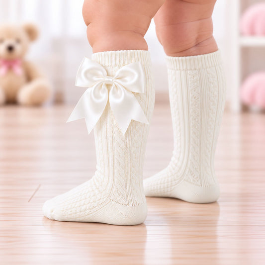 A close-up of a baby's legs wearing cream-colored, cable-knit knee-high socks with a large satin bow on the front of one sock, standing on a light wooden floor.