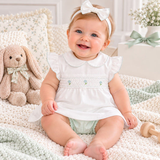 A smiling baby girl wearing a white smocked dress with ruffle sleeves, a white bow headband, and a pale green diaper, sitting on a cream knit blanket next to a plush bunny toy.