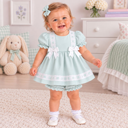 A toddler girl wearing a light mint green baby dress with white bows and trim, standing in a bright, cozy nursery room.