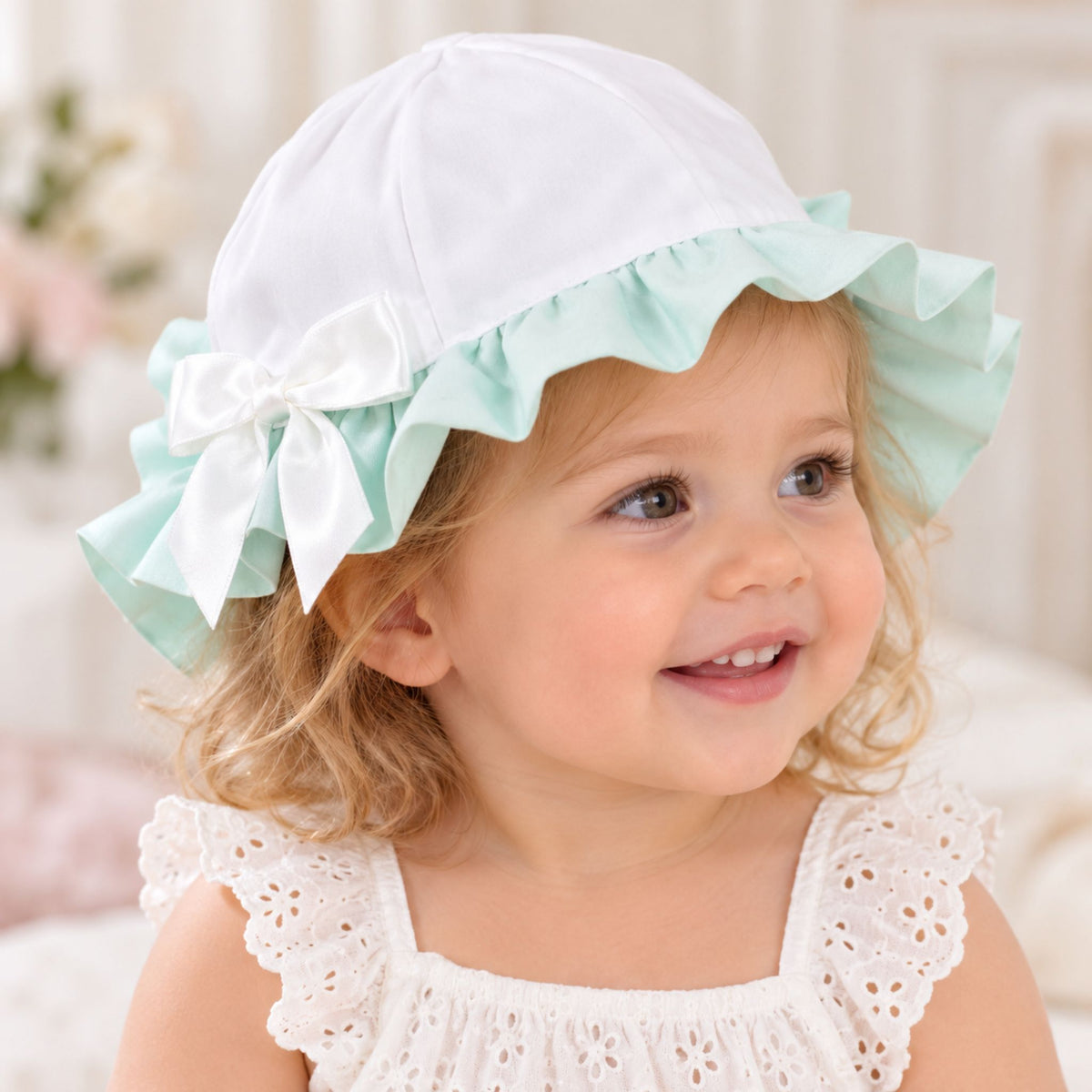 A smiling toddler girl wearing a white bonnet with a mint green ruffled brim and a white bow, paired with a white eyelet dress.