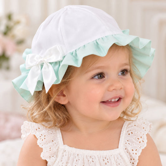 A smiling toddler girl wearing a white bonnet with a mint green ruffled brim and a white bow, paired with a white eyelet dress.