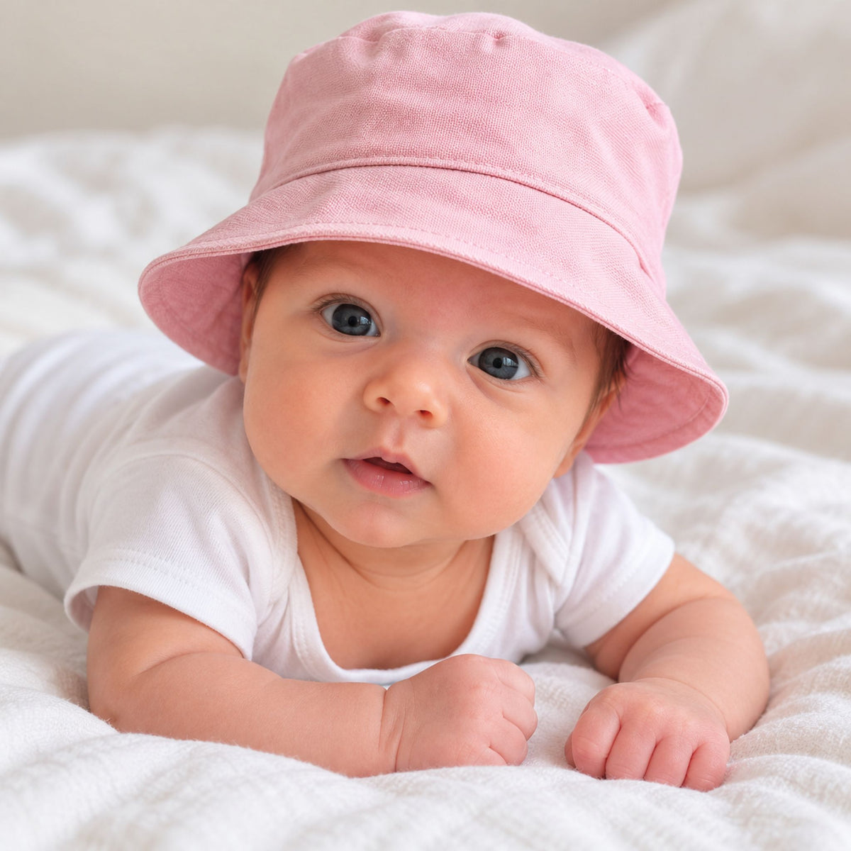 A baby lying on a white textured blanket, wearing a soft pink bucket hat and a white short-sleeve bodysuit.