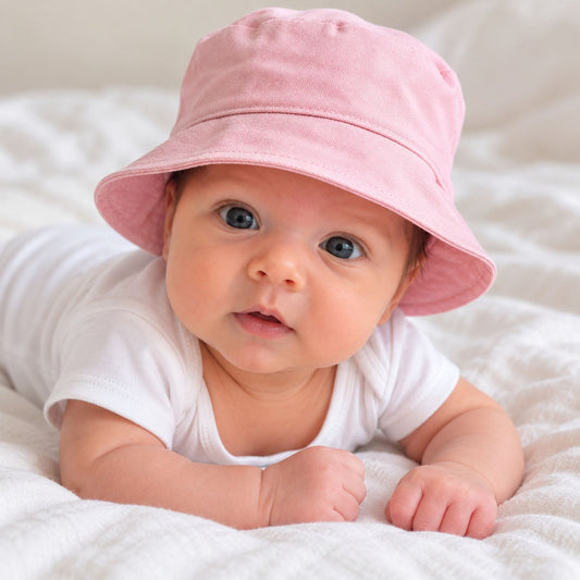 A baby lying on a white textured blanket, wearing a soft pink bucket hat and a white short-sleeve bodysuit.