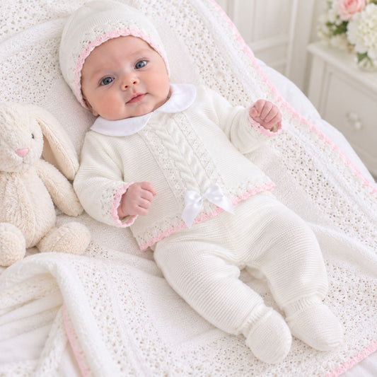 A baby wearing a white-colored knit footed suit with pink trim, a matching bonnet, and a white bow, lying on a white crocheted blanket next to a plush bunny toy.