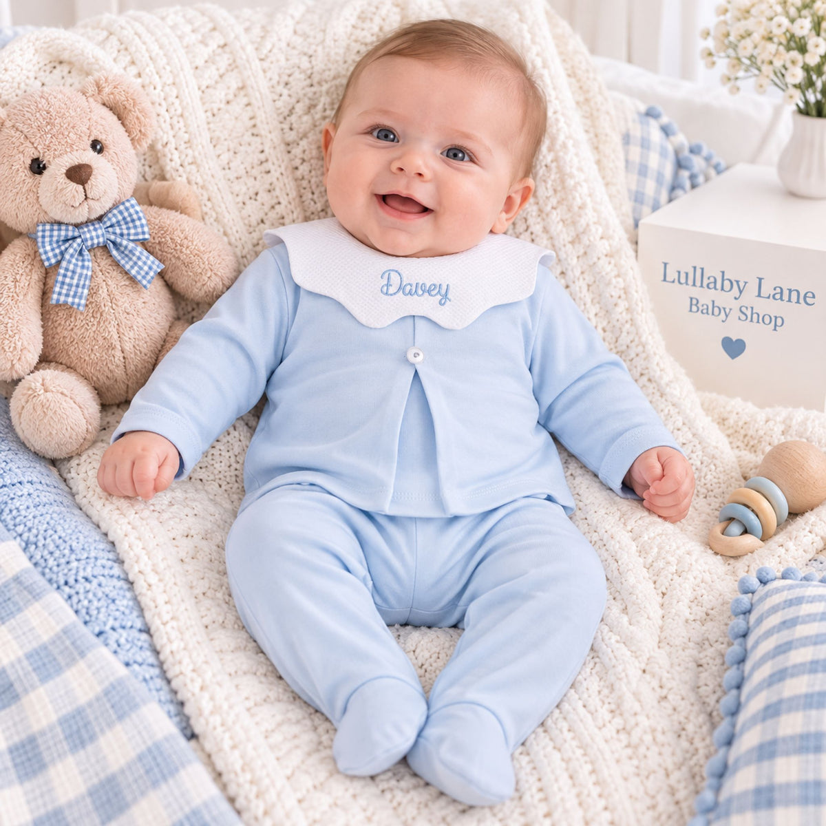 A baby wearing a light blue footed sleeper with a white bib collar embroidered with the name 'Davey' lies on a cream knit blanket, surrounded by a teddy bear, pillows, and a box reading 'Lullaby Lane Baby Shop'.