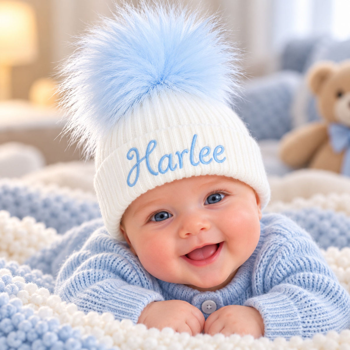 A smiling baby lying on a soft blanket, wearing a white knit pom-pom beanie with the name 'Harlee' embroidered in light blue script. The baby is also wearing a light blue knit cardigan, with a teddy bear blurred in the background.