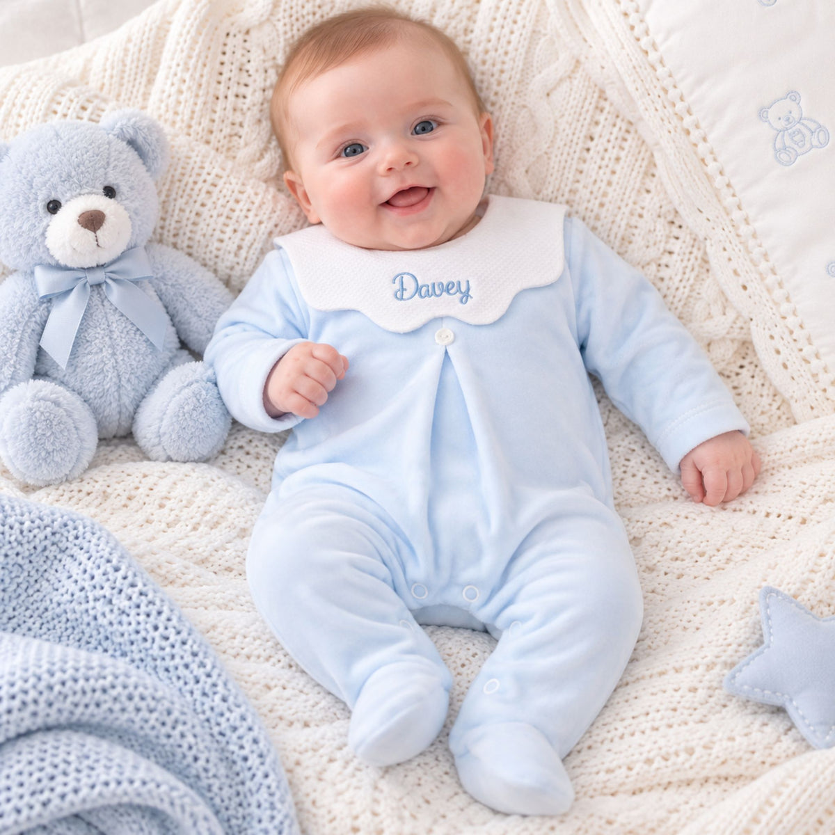 A baby lying on a cream knit blanket wearing a light blue footed sleeper with a white bib collar personalized with the name 'Davey' in blue. A light blue teddy bear sits to the left.