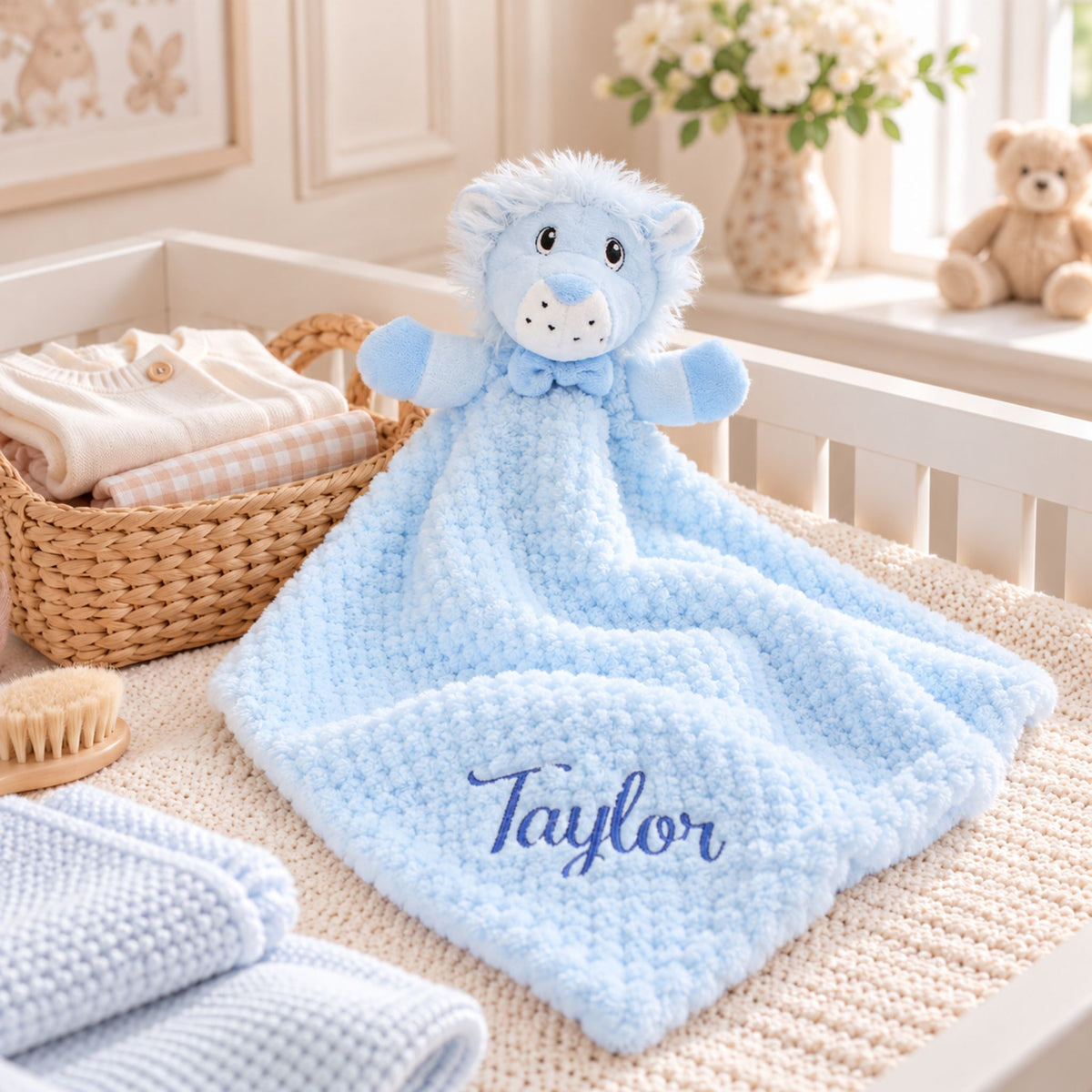 A light blue plush lion baby lovey blanket with the name 'Taylor' embroidered in blue script, displayed in a soft nursery setting next to a woven basket of folded baby clothes.