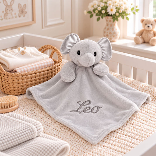 A personalized baby comforter featuring a plush elephant head and a soft gray blanket with the name 'Leo' embroidered on it, styled on a baby bed in a nursery setting.