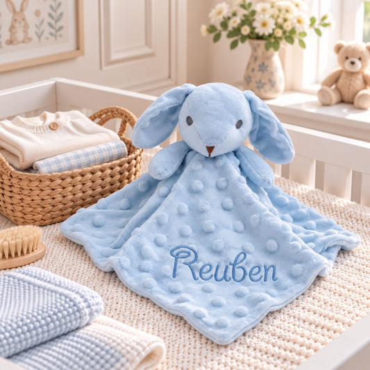 A light blue plush bunny lovey blanket with a textured bubble surface and the name 'Reuben' embroidered in blue on the front, styled in a baby nursery setting.