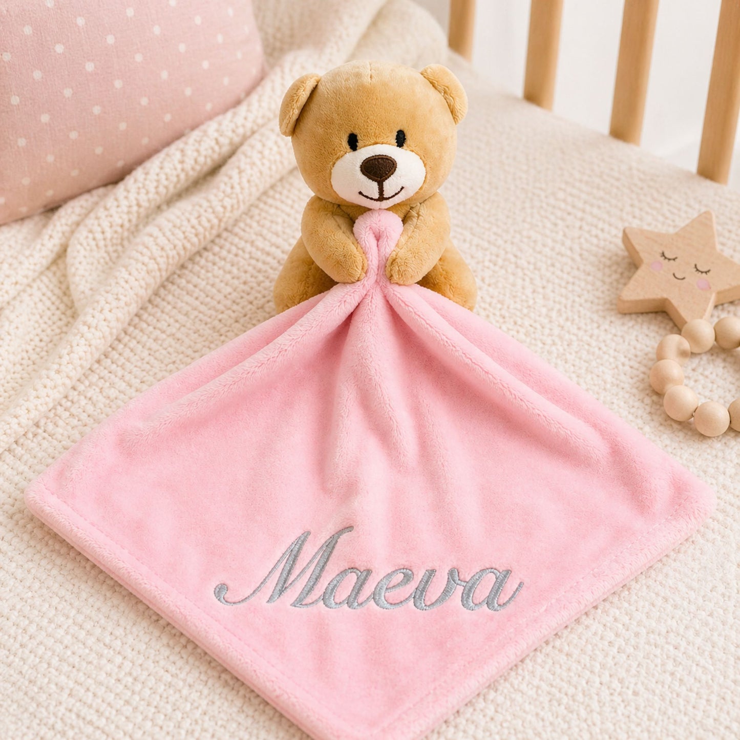 Teddy bear holding a pink blanket with 'Maeva' embroidered on it, placed on a crib with star-shaped wooden toy.