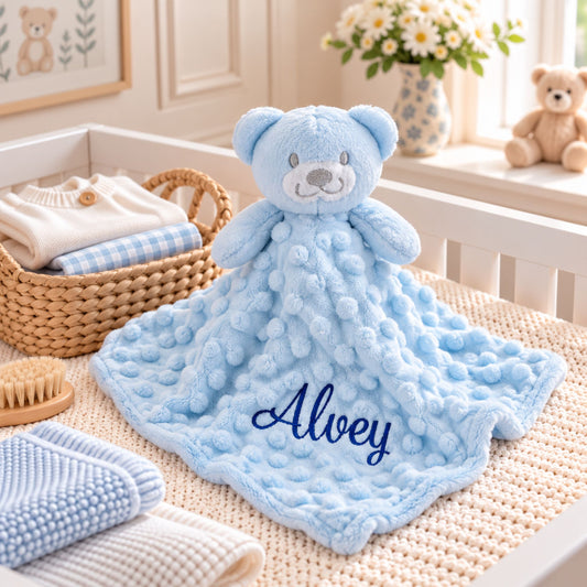 A personalized baby security blanket featuring a plush light blue teddy bear and a textured bubble blanket, embroidered with the name 'Alway' in navy blue. The blanket is styled in a soft, neutral nursery setting next to a woven basket of folded clothes.