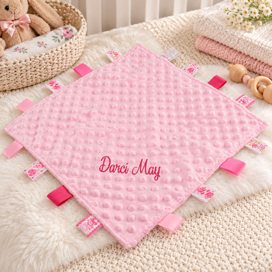 A pink textured baby lovey blanket with 'Darci May' embroidered on the front, surrounded by pink and floral jute tabs, styled on a white faux fur blanket with baby accessories.