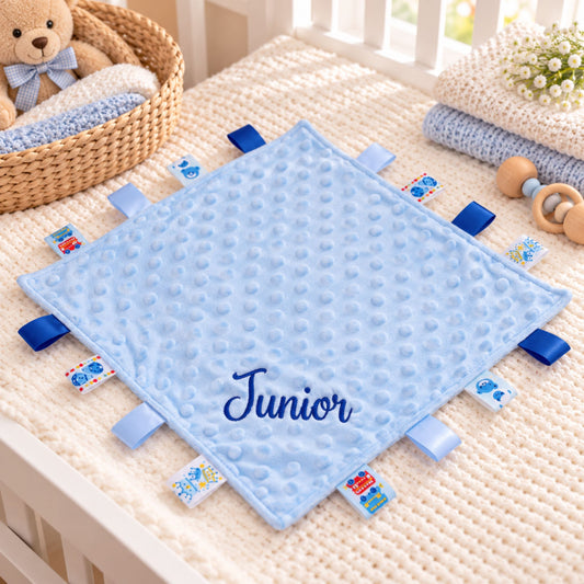 A personalized baby lovey blanket in light blue with a raised bumpy texture, featuring the name 'Junior' embroidered in navy blue on the front. The blanket is decorated with multiple blue fabric tabs on the edges, each printed with a different cartoon character.