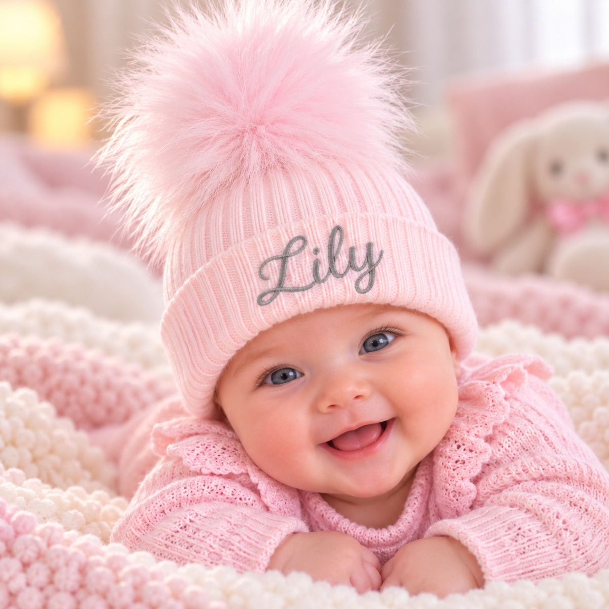 A smiling baby wearing a pink knit pom-pom beanie with the name 'Lily' embroidered on the front, lying on a pink and cream textured blanket.