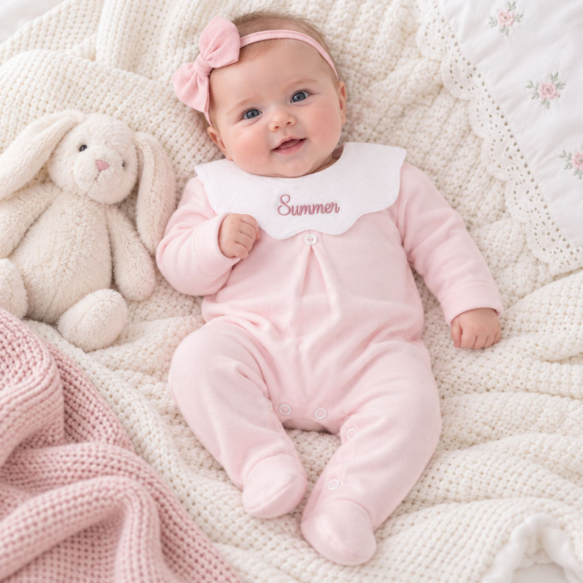 A baby lying on a cream knit blanket with a plush bunny toy, wearing a light pink footed onesie with a white bib that says 'Summer' in pink script, a pink bow headband, and a pink bonnet.
