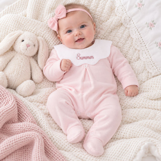 A baby lying on a cream knit blanket with a plush bunny toy, wearing a light pink footed onesie with a white bib that says 'Summer' in pink script, a pink bow headband, and a pink bonnet.