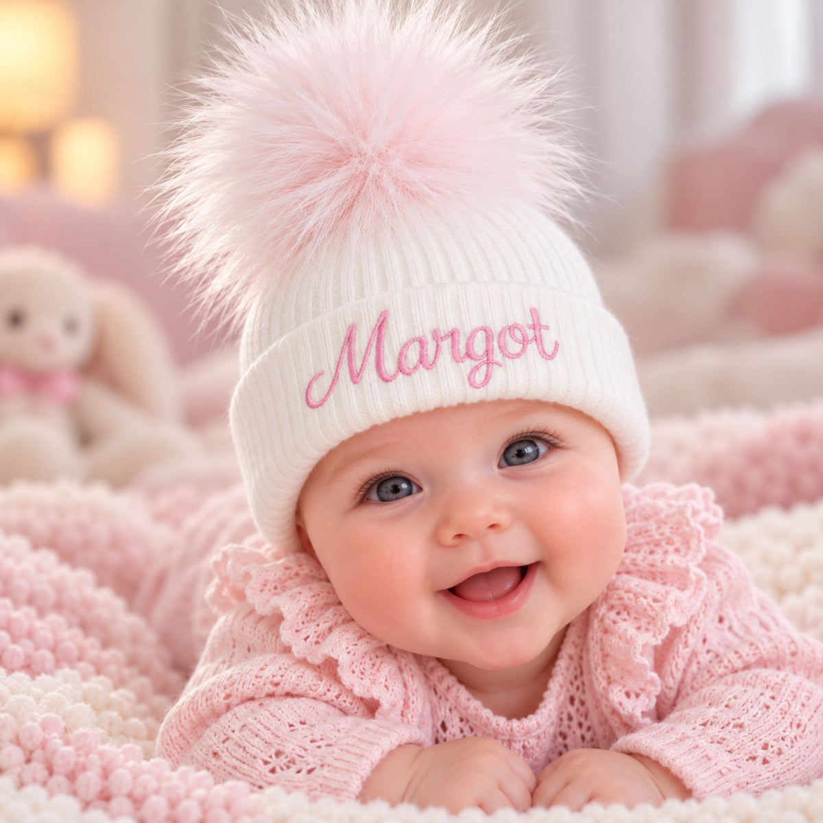 A smiling baby wearing a white knit beanie with a large fluffy pink pom-pom and the name 'Margot' embroidered in pink script on the front, posed on a pink knitted blanket.