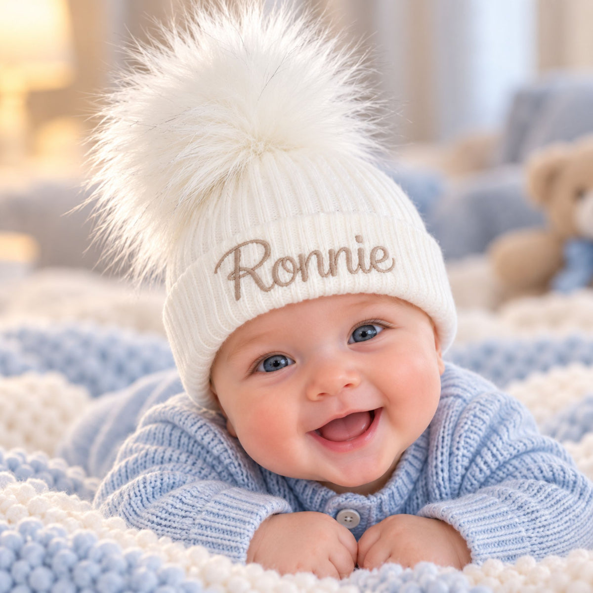 A smiling baby lying on a blue and cream knit blanket, wearing a white ribbed knit pom-pom beanie with the name 'Ronnie' embroidered in brown on the front.