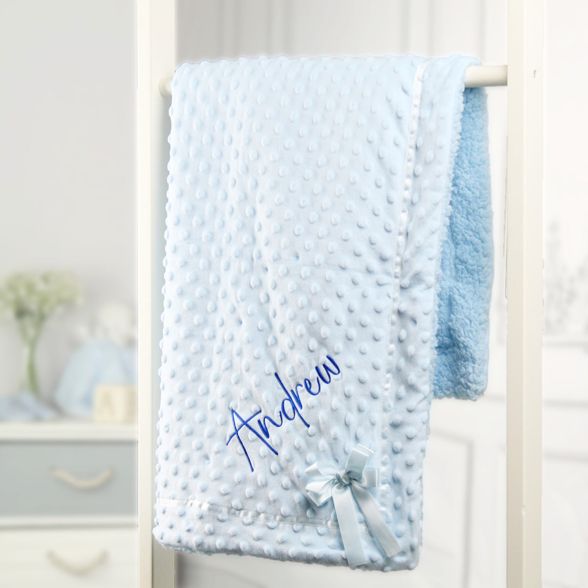 A light blue baby blanket with a textured dotty pattern, personalized with the name 'Andrew' in blue embroidery and finished with a matching ribbon bow, hanging on a white wooden stand.