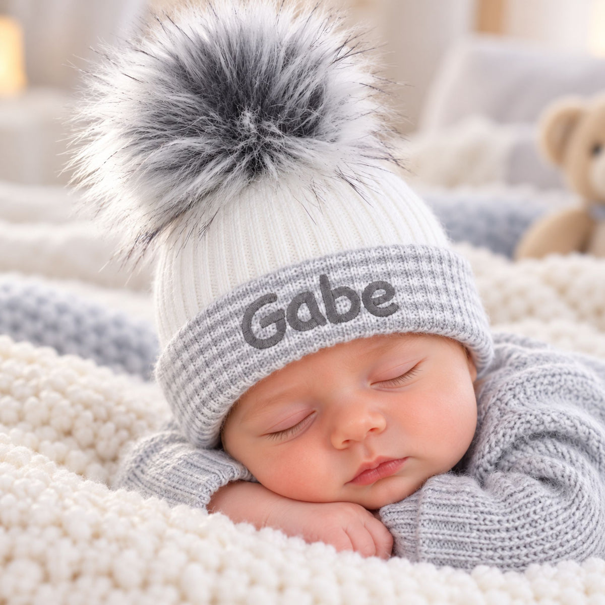A sleeping newborn baby wearing a white and gray knit beanie with a large gray-and-white pom-pom and the name 'Gabe' embroidered on the front, resting on a soft cream knit blanket.