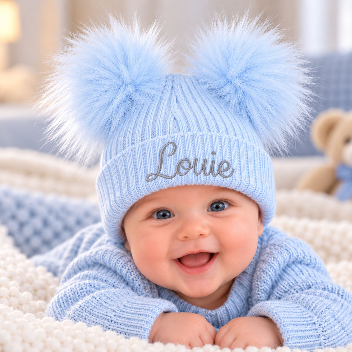 A smiling baby wearing a light blue ribbed knit beanie with two fluffy pom-poms and the name 'Louie' embroidered on the front, lying on a soft blanket.