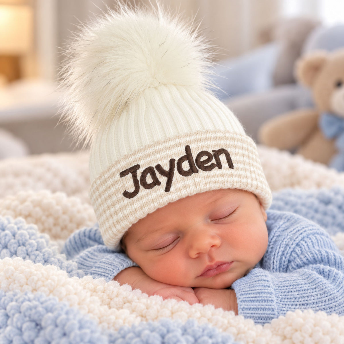 A sleeping newborn baby wearing a cream beige ribbed knit pom-pom beanie with the name 'Jayden' embroidered in brown on the front, nestled in a blue and cream textured blanket.