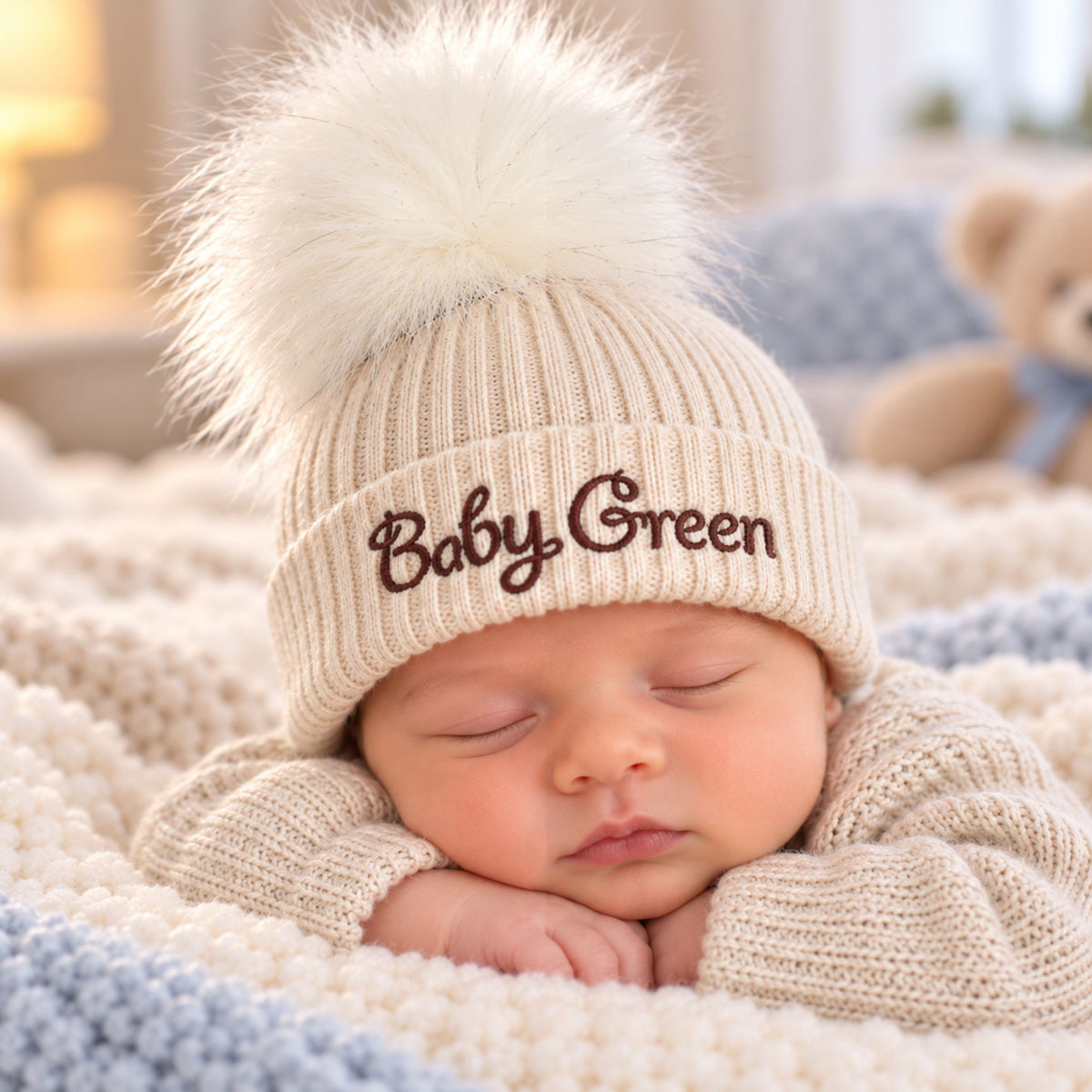 A sleeping newborn baby wearing a beige ribbed knit pom-pom beanie with 'Baby Green' embroidered in brown on the front, resting on a soft cream and blue knit blanket.