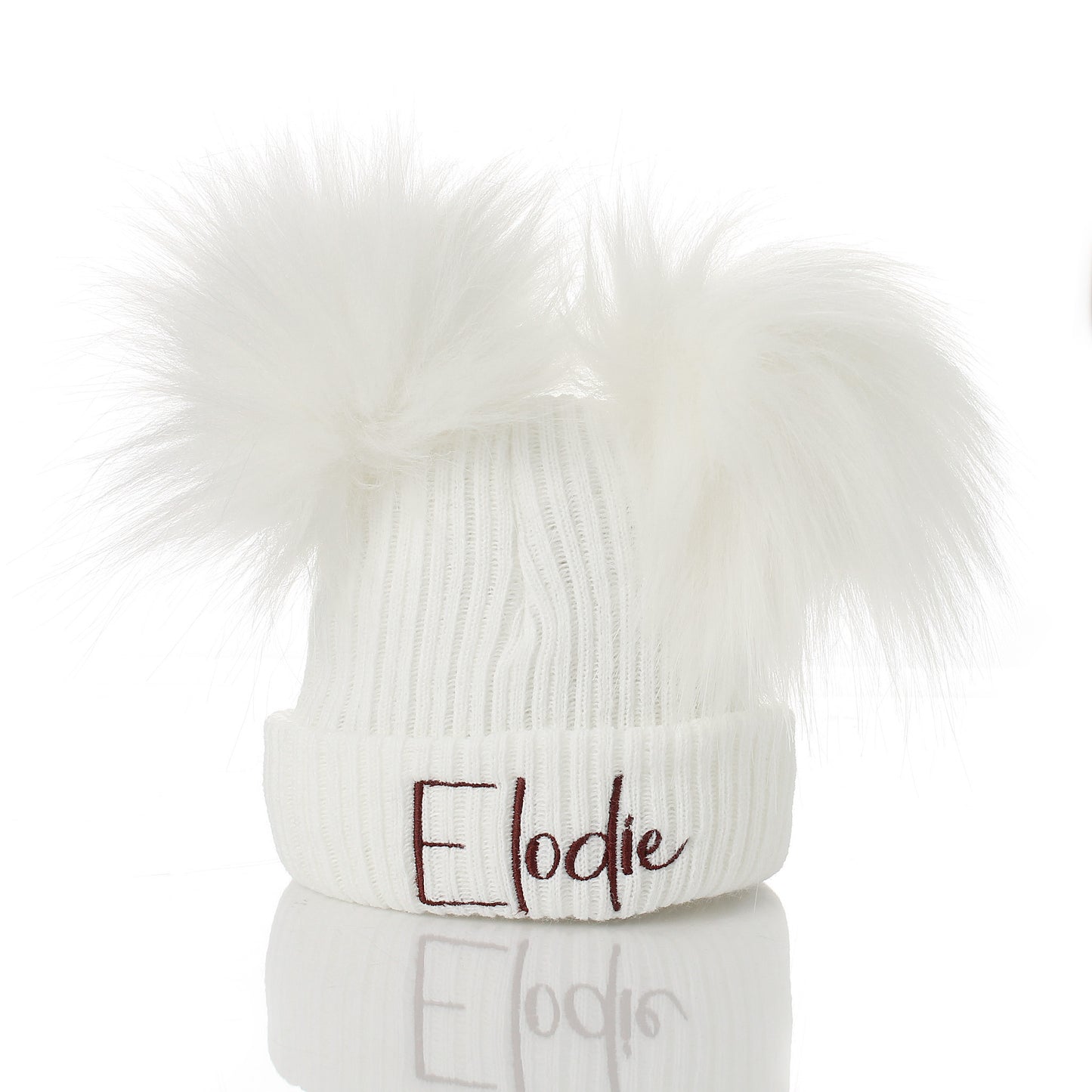 A white ribbed knit beanie hat with two fluffy faux-fur pom-poms on top and the name 'Elodie' embroidered in dark brown script on the front cuff, shown on a white background with a reflection below.