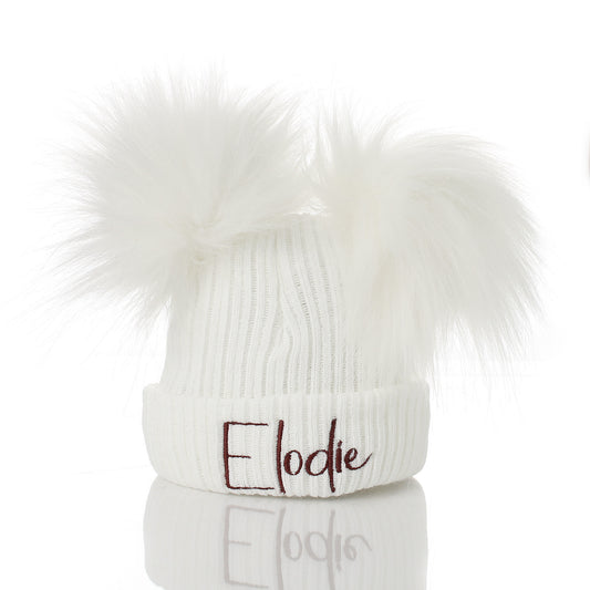 A white ribbed knit beanie hat with two fluffy faux-fur pom-poms on top and the name 'Elodie' embroidered in dark brown script on the front cuff, shown on a white background with a reflection below.