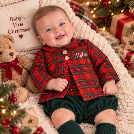 A baby lying on a cream knit blanket, wearing a red and green plaid long-sleeve shirt with wooden buttons and dark green bloomers. The scene is decorated for Christmas with a teddy bear, a pillow that says 'Baby's First Christmas', wrapped gifts, and a tree with lights.