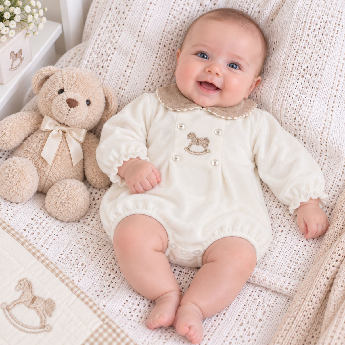 A smiling baby wearing a cream-colored long-sleeve romper with a beige ruffled collar and a small rocking horse applique, lying on a textured cream blanket next to a plush teddy bear.